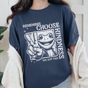 Op de afbeelding: Marineblauw T-shirt met een witte afbeelding van een kikker die een vredesteken maakt. De tekst op het shirt luidt "Remember Choose Kindness, You Got This."