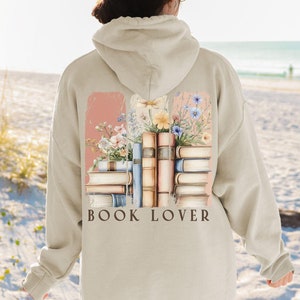 Op de afbeelding: Een beige hoodie met een afbeelding van een stapel boeken en bloemen. De tekst "BOOK LOVER" is onder de afbeelding gedrukt.