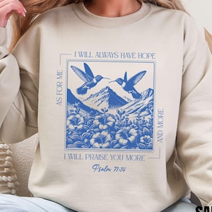 Op de afbeelding: Zandkleurige sweatshirt met een blauw vierkant ontwerp met twee kolibries, bergen en bloemelementen. De tekst luidt "I WILL ALWAYS HAVE HOPE" en "I WILL PRAISE YOU MORE" met "Psalm 71:14". Het woord "SAND" staat onderaan.