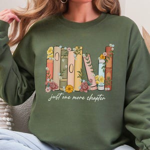 Op de afbeelding: Een groen sweatshirt met een afbeelding van een stapel boeken met bloemen en de tekst "Just one more chapter".