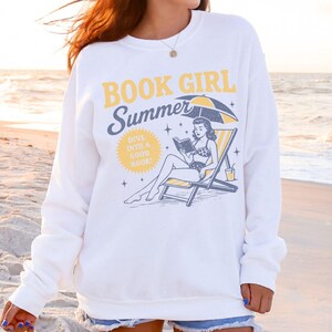 Op de afbeelding: Witte sweatshirt met de tekst "BOOK GIRL Summer" en een afbeelding van een vrouw die op een strandstoel onder een parasol leest. De sweatshirt heeft ook de tekst "DIVE INTO A GOOD BOOK!"