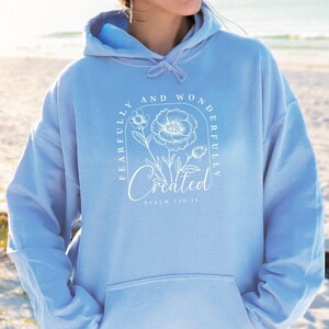 Könnte beinhalten: Hellblauer Hoodie mit einem weißen Grafikdesign, das eine Blume und den Text "Fearfully and Wonderfully Created" mit dem Bibelvers "Psalm 139:14" zeigt.