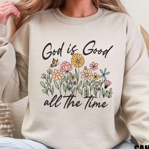 Könnte beinhalten: Ein beiger Pullover mit dem Aufdruck "God is Good all the Time" auf der Vorderseite. Der Text ist von einem Blumendesign mit Schmetterlingen umgeben.