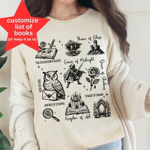 Op de afbeelding: Crèmekleurig grafisch T-shirt met illustraties van boeken en titels, waaronder "Throne of Glass" en "The Assassin's Blade". Het shirt heeft ook de tekst "customize list of books (or keep it as is)" in een rode ster en het woord "NATURAL" onderaan.