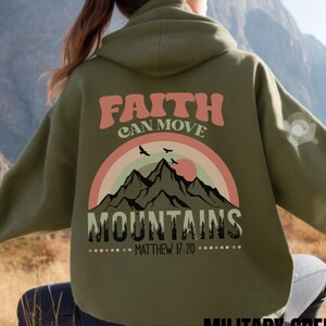 Op de afbeelding: Een olijfgroene hoodie met een graphic op de achterkant met de tekst "FAITH CAN MOVE MOUNTAINS" en "MATTHEW 17:20". Het ontwerp bevat een veelkleurige boogvormige regenboog, een silhouet van een bergketen, dennenbomen en vogels.