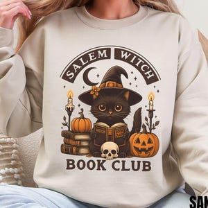 Puede incluir: Una sudadera de cuello redondo color arena con un gráfico de un gato negro con sombrero de bruja, leyendo un libro titulado Witch Book Club. El diseño incluye calabazas, velas, una calavera y el texto Salem Witch Book Club en marrón.