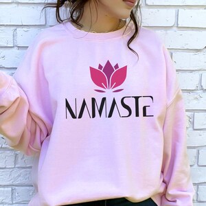 Puede incluir: Una sudadera rosa claro con la palabra "NAMASTE" en fuente negra debajo de un diseño de flor de loto magenta. La palabra "PINK" está en la esquina inferior derecha. La sudadera es de estilo casual.