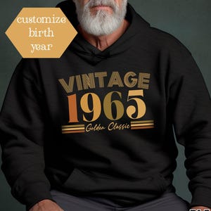 Può includere: Felpa con cappuccio nera con la scritta "VINTAGE 1965 Golden Classic" in lettere oro e arancioni. La felpa ha un cappuccio con coulisse e una tasca frontale. Il testo "customize birth year" è in un esagono dorato.
