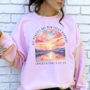 Könnte beinhalten: Rosafarbener Pullover mit einem Aquarell-Sonnenuntergangsmotiv und dem Text "His mercies are new every morning" und "Lamentations 3:22-23".