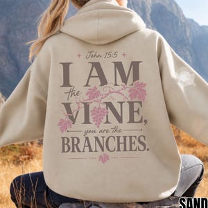 Può includere: Una felpa con cappuccio color sabbia con una scritta marrone scuro sul retro che recita John 15:5 I AM the VINE, you are the BRANCHES. Il design include illustrazioni rosa di viti e foglie integrate nella scritta.