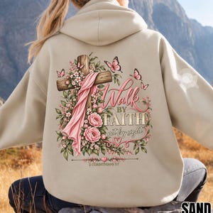 Könnte beinhalten: Ein sandfarbener Kapuzenpullover mit einem Rückendruck, der ein Holzkreuz zeigt, das mit rosa Stoff drapiert und von rosa Blumen und Schmetterlingen umgeben ist. Der Text lautet "Walk by faith not by sight" und "2 Corinthians 5:7" in Schreibschrift.