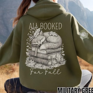 Op de afbeelding: Een olijfgroene hoodie met een afbeelding op de achterkant. Het ontwerp toont een vos die slaapt op een stapel boeken, omringd door herfstbladeren. De tekst ALL BOOKED For Fall staat in witte letters in een combinatie van stijlen.