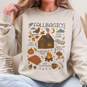 Op de afbeelding: Beige sweatshirt met een grafisch ontwerp met de tekst "#FALLBASICS" en herfstthema-illustraties. Het ontwerp bevat een hut, pompoen, kampvuur en de woorden "Get Cozy", "Stay Home" en "Read a Book".