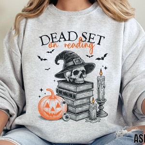 Op de afbeelding: Een lichtgrijze sweater met ronde hals en een grafische print met de tekst "DEAD SET on reading". Het ontwerp bevat een schedel met een heksenhoed op een stapel boeken, een oranje pompoen, twee brandende kaarsen en kleine vleermuizen.