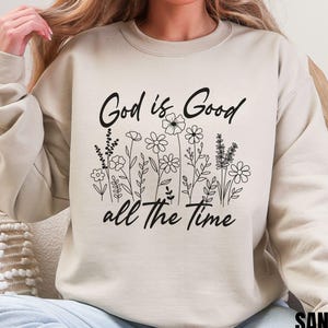 Könnte beinhalten: Ein beiger Pullover mit dem Aufdruck "God is Good all the Time" in Schwarz. Der Text ist von einem schwarzen Umriss von Blumen umgeben.
