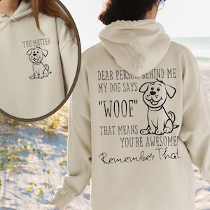 Op de afbeelding: Een beige hoodie met een zwarte hondenillustratie en de tekst "You Matter" op de voorkant. De achterkant van de hoodie heeft een zwarte hondenillustratie en de tekst "Dear Person Behind Me My Dog Says "Woof" That Means You're Awesome! Remember That".