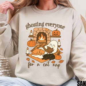 Op de afbeelding: Zandkleurige sweatshirt met een afbeelding van een open haard, pompoenen, een spook en slapende katten. De tekst luidt "ghosting everyone for a cat nap". Het woord "SAND" staat onderaan gedrukt.