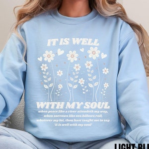 Könnte beinhalten: Hellblauer Pullover mit dem Text "IT IS WELL WITH MY SOUL" und einem Blumenmuster. Der Text unter dem Blumenmuster lautet: "when peace like a river attendeth my way, when sorrows like sea billows roll, whatever my lot, thou hast taught me to say 'it is well with my soul'".