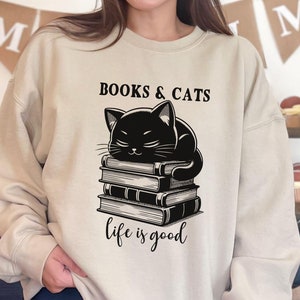 Könnte beinhalten: Ein beiger Pullover mit einer schwarzen Katze, die auf einem Stapel Bücher schläft. Der Text "BOOKS & CATS life is good" ist auf der Vorderseite des Pullovers aufgedruckt.