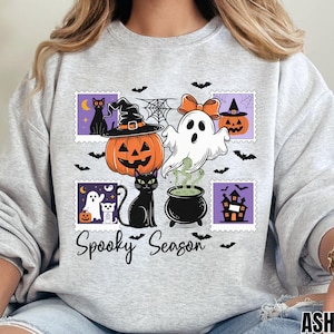 Op de afbeelding: Lichtgrijze sweatshirt met een Halloween-thema. Het ontwerp toont een pompoen met een heksenhoed, een spook, een zwarte kat en de tekst "Spooky Season". Vleermuizen en spinnenwebben maken het ontwerp compleet.