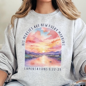 Könnte beinhalten: Hellgraues Sweatshirt mit einem Aquarelldesign eines Sonnenuntergangs über einem Fluss, mit dem Text "HIS MERCIES ARE NEW EVERY MORNING" und "LAMENTATIONS 3:22-23" in einem Halbkreis über und unter dem Bild.