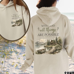 Könnte beinhalten: Ein sandfarbener Kapuzenpullover mit einem Grafikdruck von einem See, einer Holzhütte und einem Boot auf der Rückseite sowie einer kleineren Version auf der Brust. Der Text lautet "WITH GOD all things ARE POSSIBLE Matthew 19:26" in dunkler Schrift.