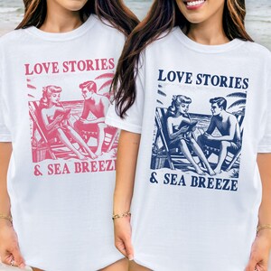Op de afbeelding: Witte T-shirts met een retro grafisch ontwerp. Het ontwerp toont een stel op een strand dat leest, met de woorden "LOVE STORIES & SEA BREEZE" in een rechthoekige doos. Eén shirt heeft een roze afbeelding, de andere een blauwe afbeelding.