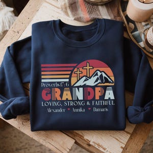 Op de afbeelding: Marineblauw sweatshirt met een grafisch ontwerp. Het ontwerp bevat het woord "GRANDPA" in oranje en rood, met de tekst "LOVING, STRONG & FAITHFUL" eronder. Het sweatshirt bevat ook de namen Alexander, Annika en Damaris.