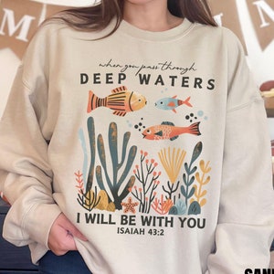 Könnte beinhalten: Beigefarbener Pullover mit einem Aufdruck von bunten Fischen, die in einem Korallenriff schwimmen. Der Text "when you pass through DEEP WATERS I WILL BE WITH YOU ISAIAH 43:2" ist auf dem Pullover aufgedruckt.