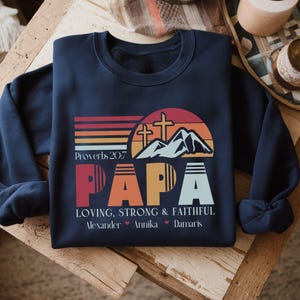Op de afbeelding: Donkerblauwe trui met een retro zonsondergang-afbeelding, bergen en kruisen. De tekst "PAPA" staat in grote, kleurrijke letters. Onder de afbeelding staat de tekst "Spreuken 20:7 LIEFHEBBEND, STERK & GETROUW" en de namen "Alexander, Annika, Damaris".
