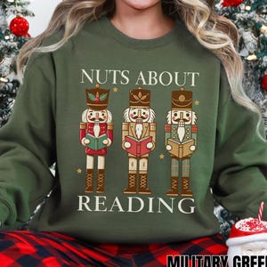 Op de afbeelding: Een militairgroen sweatshirt met de tekst "NUTS ABOUT READING" en drie geïllustreerde notenkrakerfiguren. De notenkrakers lezen boeken en dragen rode, bruine en groene outfits. De sweatshirt is een crew neck met lange mouwen.