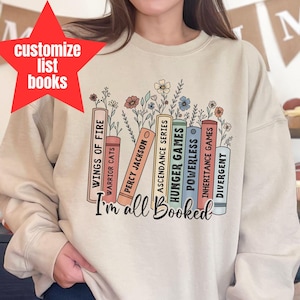 Op de afbeelding: Een licht beige sweatshirt met een afbeelding van een stapel boeken met bloemmotieven. De tekst "I'm all Booked" is onder de boeken gedrukt.