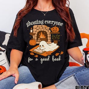 Op de afbeelding: Een zwart T-shirt met korte mouwen en een afbeelding van een geest die een boek leest voor een open haard. De tekst luidt "ghosting everyone for a good book". Het ontwerp bevat een pompoen, een lantaarn en herfstbladeren.