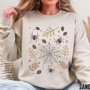 Op de afbeelding: Zandkleurige sweatshirt met een Halloween-thema. De afbeelding bevat een spinnenweb, spinnen, bladeren en bessen in bruine en beige tinten. Het woord "SAND" staat rechtsonder.