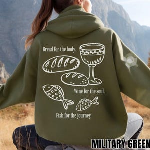 May include: Military green hoodie featuring white illustrations of bread, a wine glass, and fish. Text reads "Bread for the body," "Wine for the soul," and "Fish for the journey." The hoodie is a solid colour.