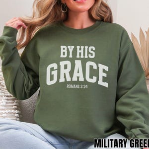 May include: Military green sweatshirt featuring the text "BY HIS GRACE" in white, with "ROMANS 3:24" underneath. The sweatshirt has a crew neck design.