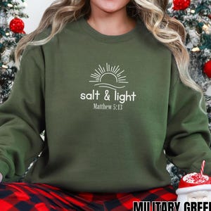 Op de afbeelding: Een legergroene sweatshirt met de witte tekst "salt & light" en "Matthew 5:13". Het ontwerp bevat een zon en een golf. De sweatshirt heeft een ronde hals.