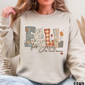 Op de afbeelding: Een zandkleurige sweater met ronde hals en het woord FALL in grote, gedessineerde blokletters met bloemen-, ruit- en bladmotieven. Daaronder staat de tekst for Jesus He never leaves in een combinatie van cursief en druk.