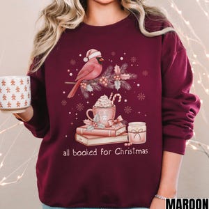 Op de afbeelding: Bordeaux sweatshirt met een kerstthema. Het ontwerp toont een kardinaal met een kerstmuts, een mok slagroom, boeken en de tekst "all booked for Christmas".