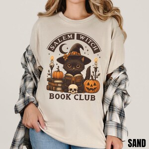 K&ouml;nnte beinhalten: Ein sandfarbenes T-Shirt mit einer Grafik einer schwarzen Katze, die einen Hexenhut tr&auml;gt und ein Buch liest. Das Design enth&auml;lt den Text Salem Witch Book Club, K&uuml;rbisse, Kerzen, eine Mondsichel und einen Totenkopf. Ein kariertes Hemd ist dar&uuml;ber geschichtet.