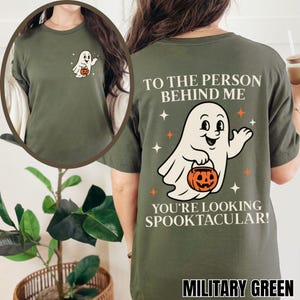 Puede incluir: Una camiseta verde oliva con un peque&ntilde;o gr&aacute;fico de un fantasma blanco en el pecho izquierdo. La parte trasera muestra un gran fantasma blanco sosteniendo una calabaza naranja, rodeado de estrellas, con el texto TO THE PERSON BEHIND ME YOU'RE LOOKING SPOOKTACULAR!