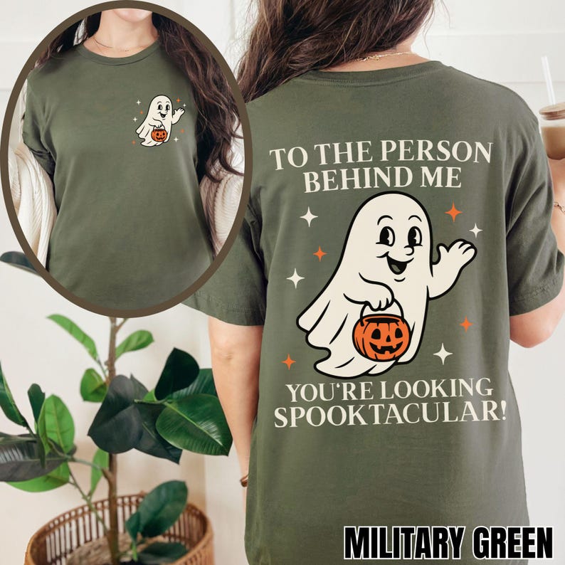 Puede incluir: Una camiseta verde oliva con un peque&ntilde;o gr&aacute;fico de un fantasma blanco en el pecho izquierdo. La parte trasera muestra un gran fantasma blanco sosteniendo una calabaza naranja, rodeado de estrellas, con el texto TO THE PERSON BEHIND ME YOU'RE LOOKING SPOOKTACULAR!