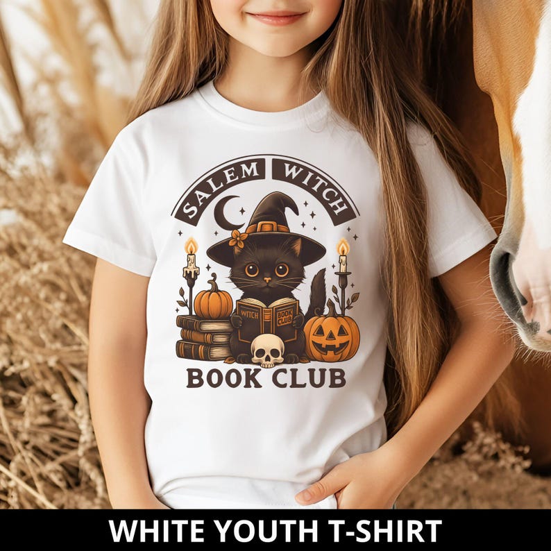 K&ouml;nnte beinhalten: Ein wei&szlig;es T-Shirt mit einer Grafik eines schwarzen Katers mit Hexenhut, der ein Buch mit dem Titel Witch Book Club liest. Das Design zeigt zwei K&uuml;rbisse, einen Totenkopf, zwei brennende Kerzen und den Schriftzug Salem Witch Book Club in Braun.