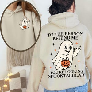 Puede incluir: Una sudadera con capucha beige que presenta un peque&ntilde;o gr&aacute;fico de fantasma en el pecho izquierdo. La parte posterior muestra un fantasma grande sosteniendo una calabaza con el texto: TO THE PERSON BEHIND ME YOU'RE LOOKING SPOOKTACULAR! y estrellas.