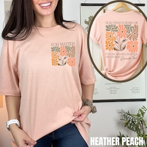 Puede incluir: Una camiseta de color melocot&oacute;n con el texto "You Matter" y un dise&ntilde;o floral. La parte posterior de la camiseta tiene el texto "Dear person behind me, you are amazing, beautiful and enough. Remember that."