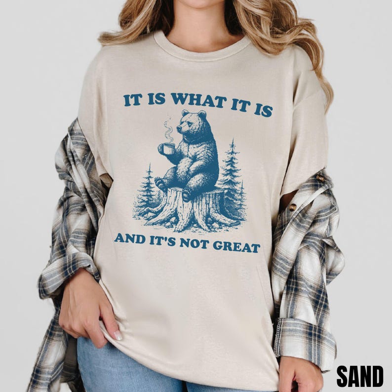 May include: Sand-colored t-shirt featuring a blue graphic of a bear sitting on a tree stump, holding a mug. The text "IT IS WHAT IT IS AND IT'S NOT GREAT" is printed above and below the bear illustration. The shirt is a casual wear item.