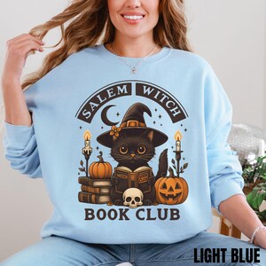 K&ouml;nnte beinhalten: Ein hellblaues Sweatshirt mit Rundhalsausschnitt und einem Grafikdruck einer schwarzen Katze mit Hexenhut, die ein Buch mit dem Titel Witch Book Club liest. Das Design umfasst einen Totenkopf, K&uuml;rbisse, Kerzen und den Text Salem Witch Book Club in Dunkelbraun.