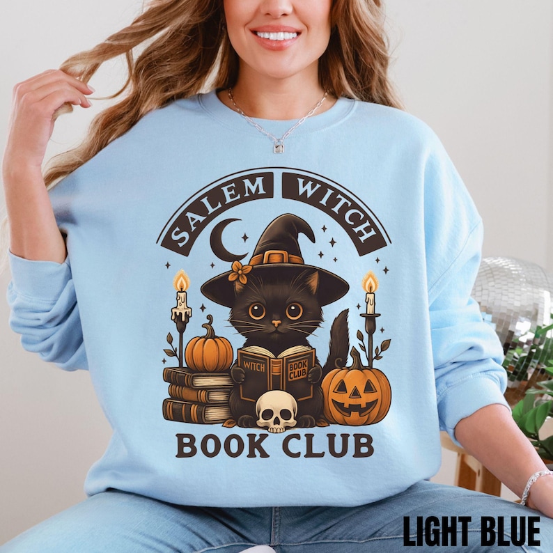 K&ouml;nnte beinhalten: Ein hellblaues Sweatshirt mit Rundhalsausschnitt und einem Grafikdruck einer schwarzen Katze mit Hexenhut, die ein Buch mit dem Titel Witch Book Club liest. Das Design umfasst einen Totenkopf, K&uuml;rbisse, Kerzen und den Text Salem Witch Book Club in Dunkelbraun.