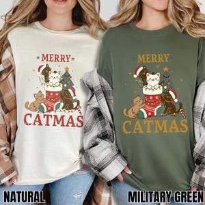 Op de afbeelding: Twee T-shirts, &eacute;&eacute;n in naturel en &eacute;&eacute;n in legergroen, tonen een "Catmas"-ontwerp. Het ontwerp bevat katten, een kerstboom en een sok. De tekst "Merry Catmas" staat boven en onder de afbeelding.
