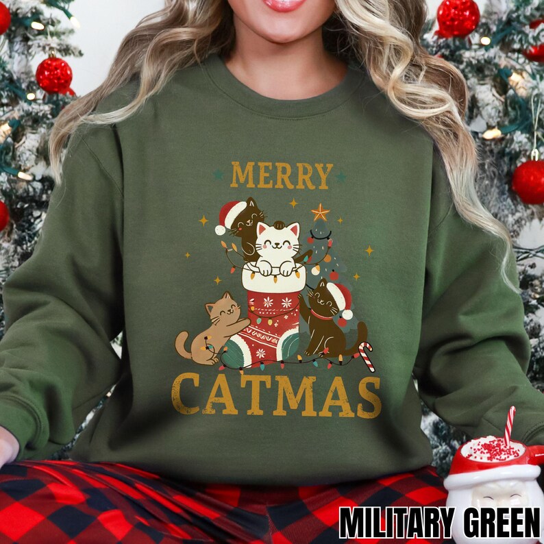 Op de afbeelding: Een legergroene sweatshirt met de woorden "MERRY CATMAS" in gouden letters. De afbeelding toont drie katten met kerstmutsen, waarvan &eacute;&eacute;n in een kerstsok. Een zuurstok en kerstverlichting zijn ook inbegrepen.